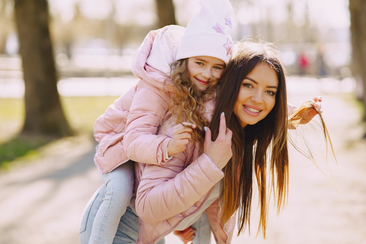 Side view of smiling little girl during piggyback ride with mother wearing warm pink jacket looking at camera while strolling in city park in sunny spring day